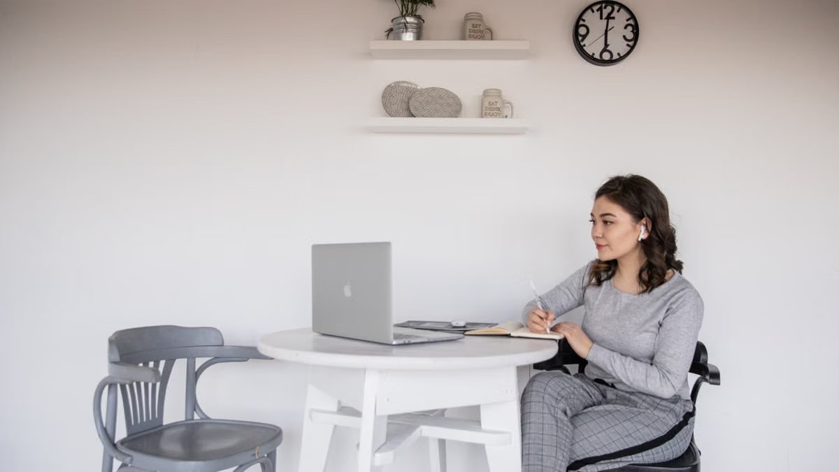 Smiling young ethnic female student in casual wear and wireless earbuds sitting with crossed legs and writing in notebook while watching netbook and studying at home